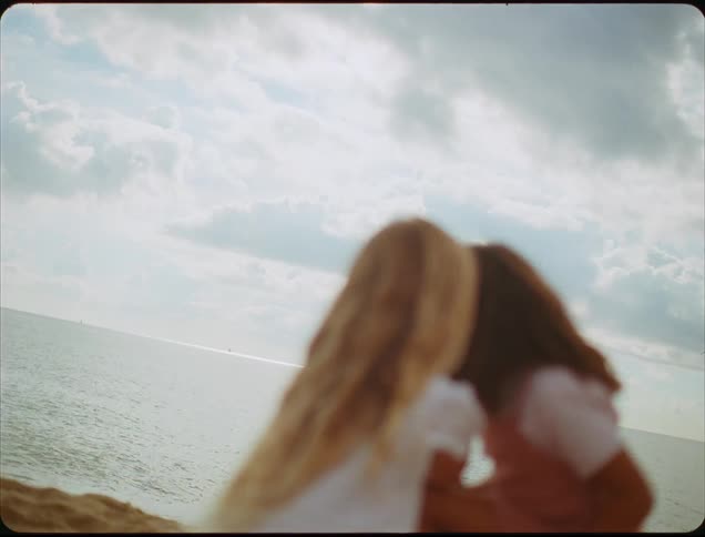 Two kids are on the beach at midday playfully chatting as one leans forward to whisper into the others ear