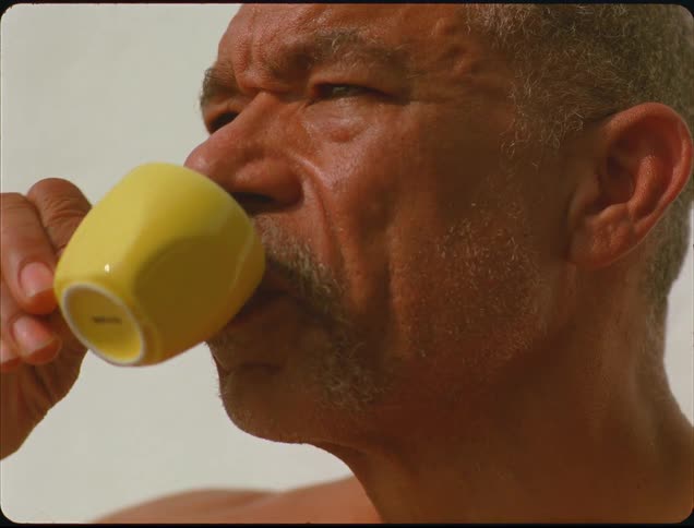 A close view of an older man with gray hair raising a small yellow cup to his lips and taking a thoughtful drink