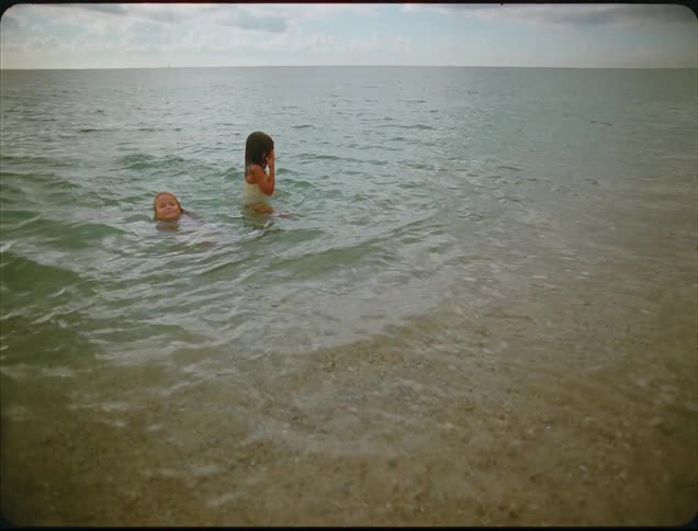 This video featuring a grainy vintage film look shows two young girls playing in the shallow cleargreen ocean water they are partially submerged facing the shore as small waves break around them under an overcast sky