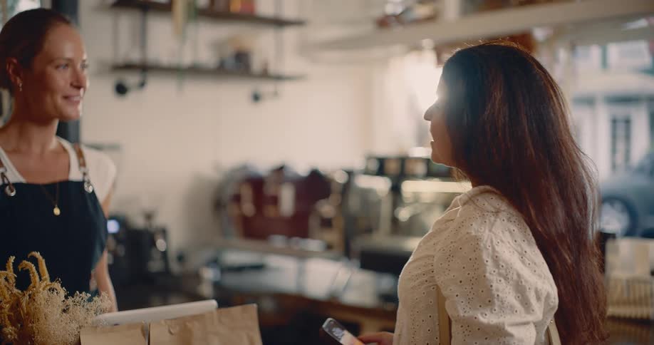 The video begins with a woman inside a coffee shop smiling and talking animatedly on her mobile phone as she approaches the counter she ends her call puts the phone down and is then greeted by a cheerful barista wearing a black apron