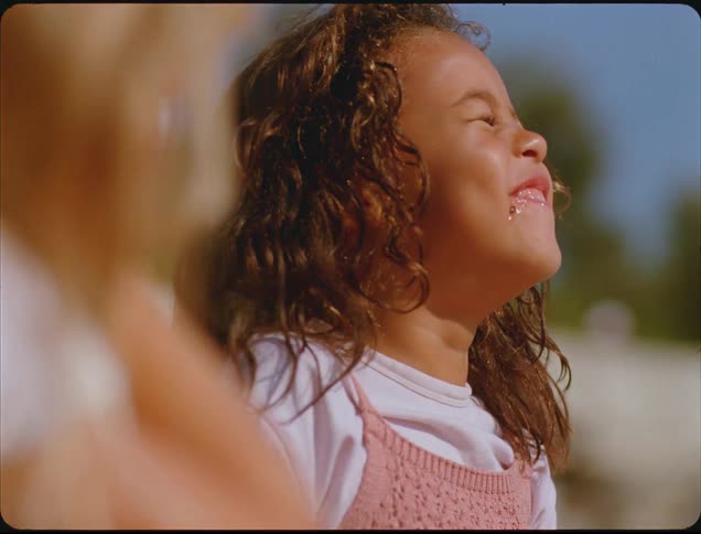 A close view of a little girl with curly brown hair eating a piece of watermelon outdoors on a sunny day