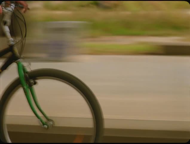 A low angle close view of a person wearing a white t shirt and shorts quickly riding a bicycle outdoors near the ocean on a bright sunny day
