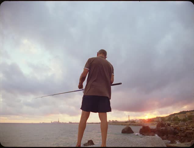 A man is standing on a rocky shoreline seen from behind and below holding a long fishing rod extending out over the water he is silhouetted against a dramatic cloudy sky with hints of a colorful sunset on the horizon over a distant cityscape