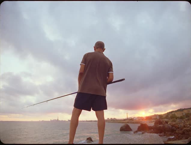 A man stands on the edge of the beach with a fishing rod in his hands and the sun rising on the horizon