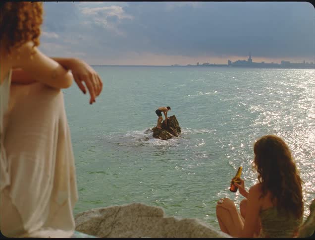 Two women are sitting on rocky terrain overlooking the sea while a man stands on a small rock formation further out in the water preparing to dive in the distance a cityscape is visible under a cloudy sky suggesting a moment of leisure near an urban a