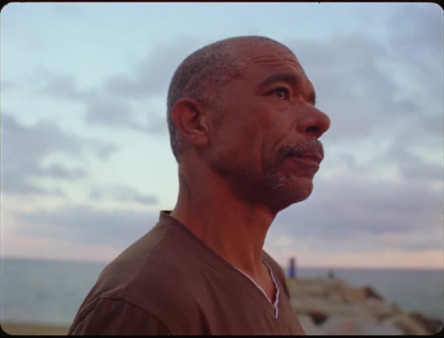A close side view of an older man with a shaved head and brown shirt standing outdoors and looking up at a cloudy sunset sky above the ocean