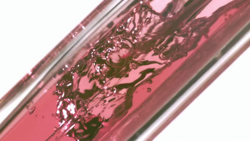 A closeup angled shot shows a viscous pink liquid being poured into a transparent glass container creating turbulent bubbling and splashing against the inner surface the reflections on the glass and the deep color of the fluid give the scene a vibran
