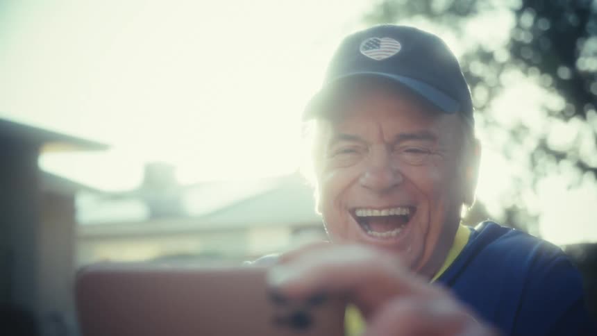 The camera is initially blinded by intense sunlight before panning down to reveal a joyful older man wearing a dark baseball cap with a us flag patch and a blue and yellow athletic shirt he is laughing exuberantly while holding an object or package just