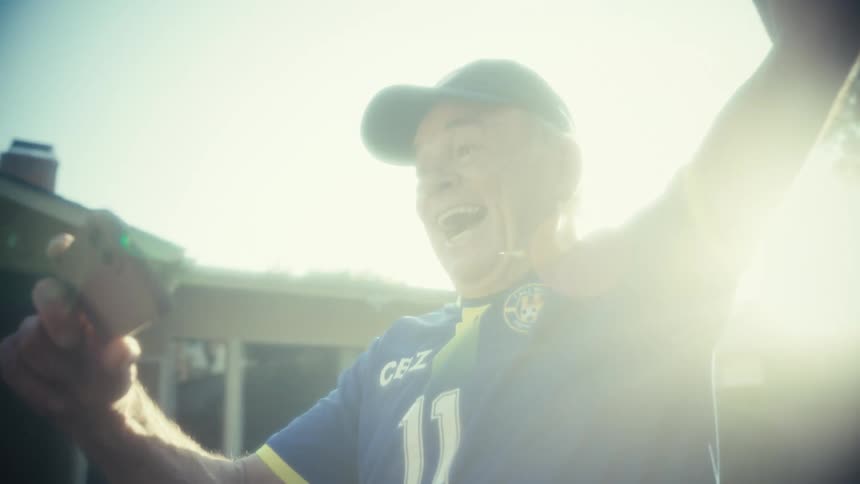 A man wearing a sports jersey intensely celebrates and smiles broadly into a handheld smartphone with brilliant sunlight flaring brightly behind him