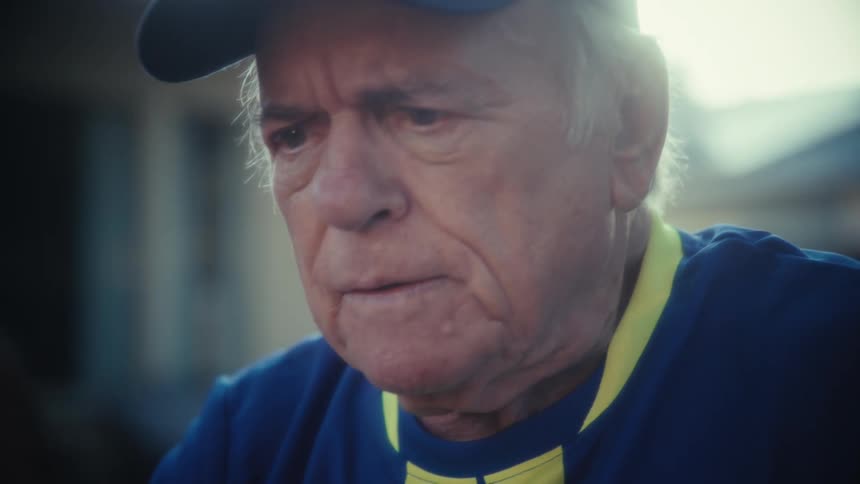The video shows a closeup of an elderly man wearing a blue and yellow sports jersey and a baseball cap he appears to be talking or reacting with an emotional expression possibly one of slight distress or frustration as his mouth is open and his brow i