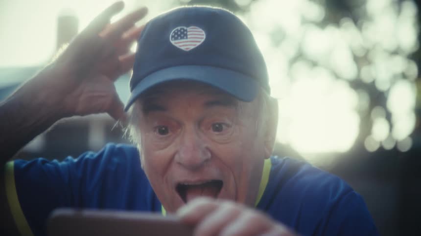 An elderly man sits outdoors in his backyard at midday wearing a hat and watching a sports event on his phone suddenly bursting with excitement