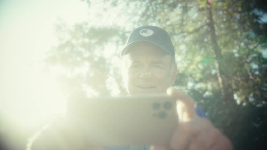 An older man wearing a baseball cap and blue and yellow shirt holds a smartphone to record or take a selfie on a bright sunny day outdoors