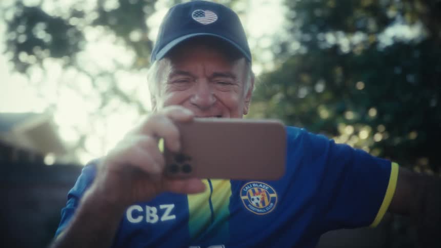 An excited man wearing a blue jersey and cap cheers loudly and pumps his fist in the air while recording a video on his phone