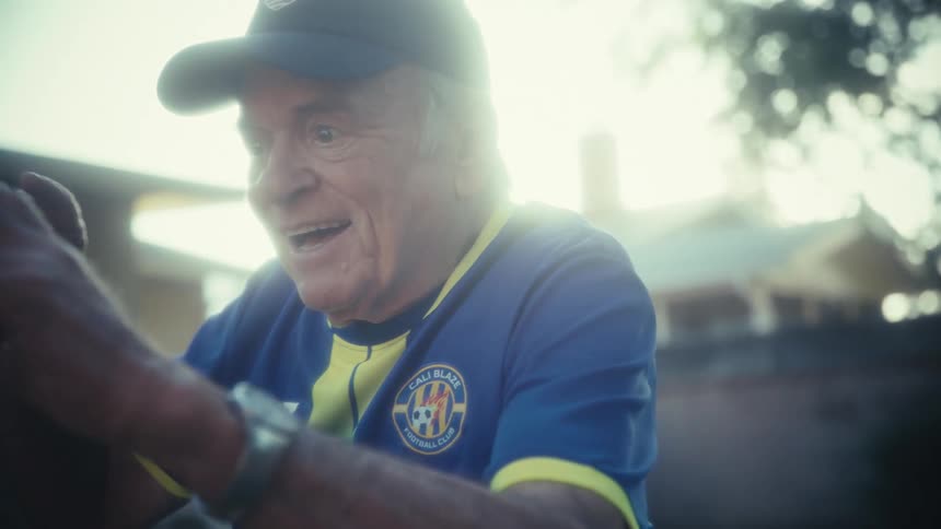 An elderly man wearing a blue and yellow soccer jersey and a baseball cap is initially seen concentrating on his phone seemingly recording or watching somethinghis expression dramatically shifts to one of excitement and shock culminating in a widemo
