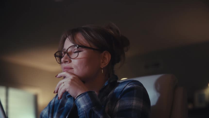 The video shows a closeup of a person with glasses wearing a plaid shirt working on a tablet or laptop in a dimly lit room they appear to be concentrating intently at first then touch their forehead or eyes suggesting fatigue or strain from work