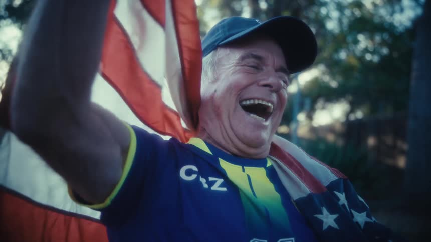 An elderly man wearing a hat and a sports jersey stands in his backyard at midday with an american flag draped over his shoulder he watches a game on his phone and suddenly bursts with excitement