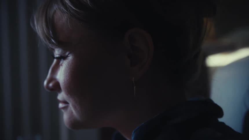 A woman holds her glasses and gazes thoughtfully before resting her hand on her chin in a dimly lit room