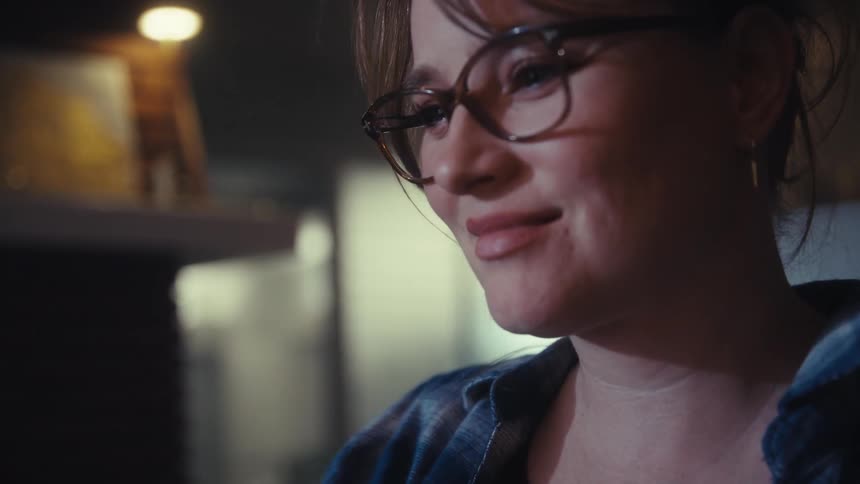 A closeup shot of a young woman smiling slightly while looking offcamera she is wearing large round eyeglasses and a plaid shirt in dimly lit indoor lighting