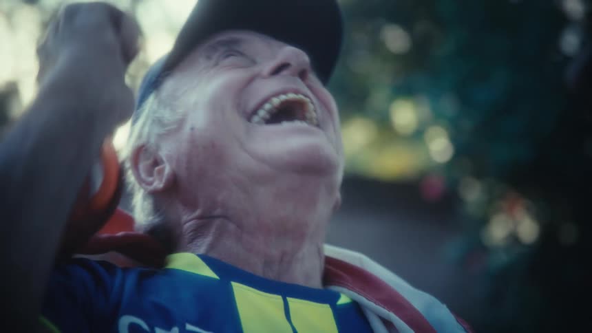 An excited man wearing a baseball cap and a sports jersey cheers loudly while smiling broadly with an american flag draped over his shoulders