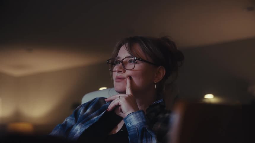 A woman wearing glasses and a plaid shirt works at night on a laptop or tablet touching her face and thinking in a warm lit interior