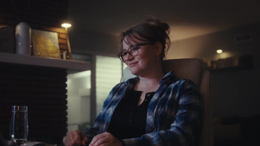 A woman with her hair up and wearing glasses and a plaid shirt is typing on a keyboard in a dimly lit room with warm interior lighting behind her