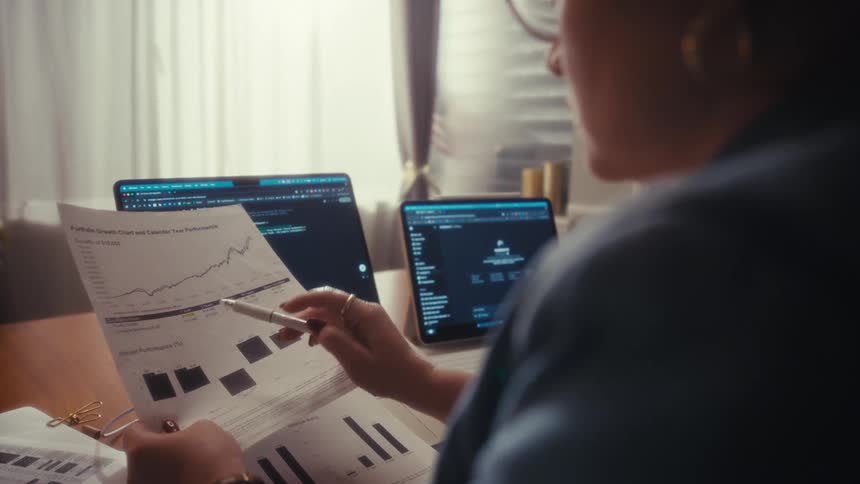 A close view over a womans shoulder shows her using a pen to analyze a printed stock chart while working between a laptop and a tablet in a sunny office