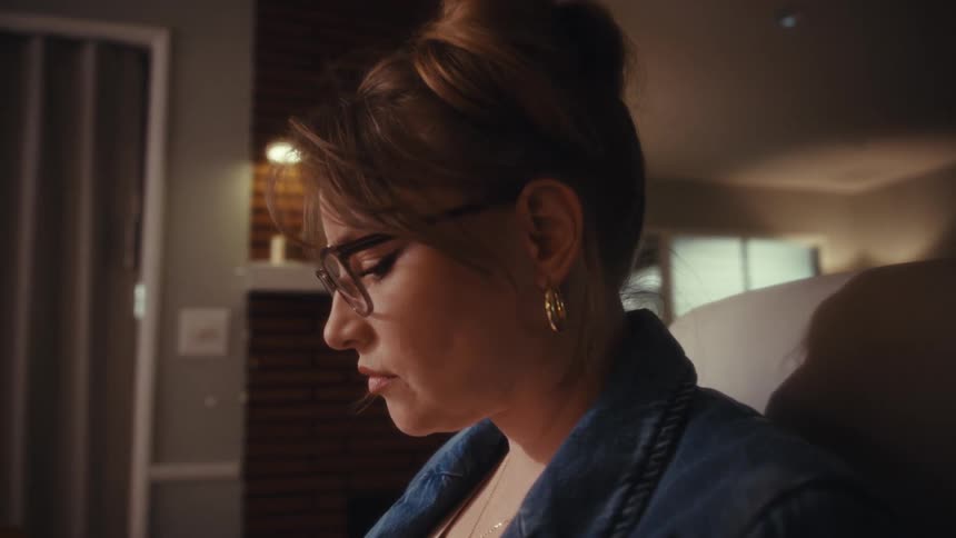 A focused woman wearing a denim jacket and gold earrings works with a stylus on a keyboard next to a laptop in a warm dimly lit home