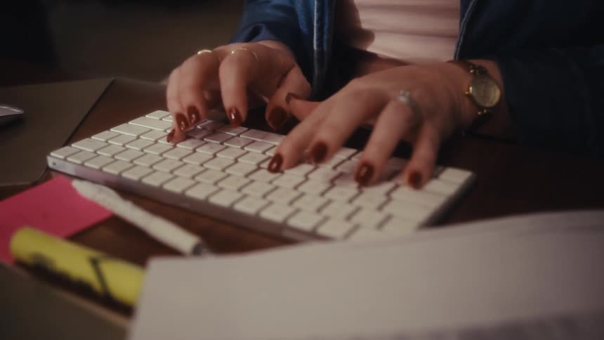 A close view shows a smiling woman then focuses on her hands with dark nails quickly typing on a white computer keyboard in a dimly lit office