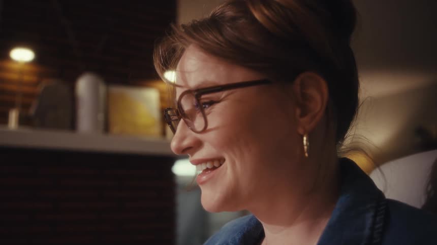 A woman with glasses laughs joyfully while looking off camera in a cozy warmly lit room