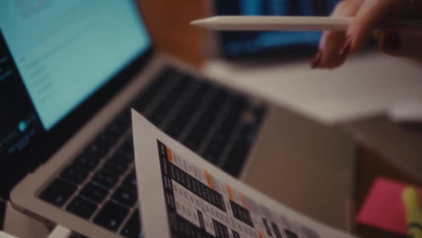 A close view focuses first on a hand analyzing a printed chart with a stylus then shifts to a laptop screen displaying a digital financial dashboard