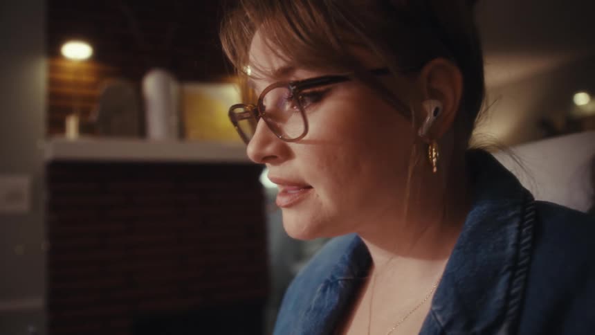 A close view shows a woman wearing a denim jacket and glasses talking on a video call while analyzing financial charts visible on her laptop screen