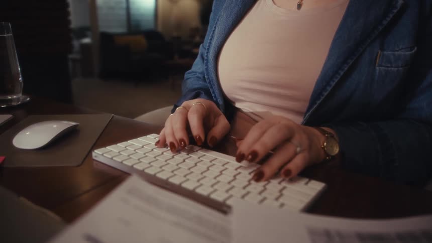 A woman wearing a denim jacket types on a white computer keyboard and uses a mouse at a wooden desk in a dimly lit home office