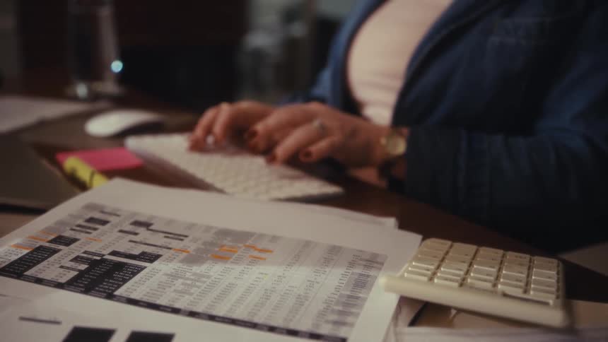Paper charts and graphs are scattered on a work desk while a woman types on a computer keyboard