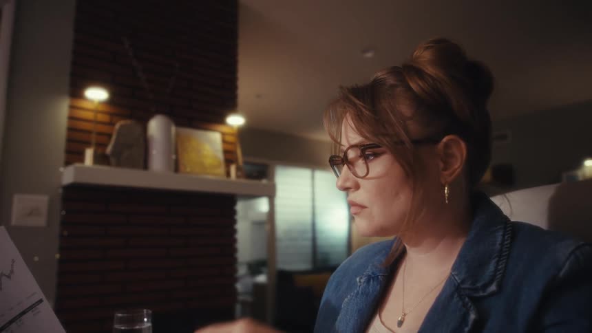 A close view shows a woman wearing glasses and a denim jacket seriously studying a document containing charts and graphs in a dimly lit room