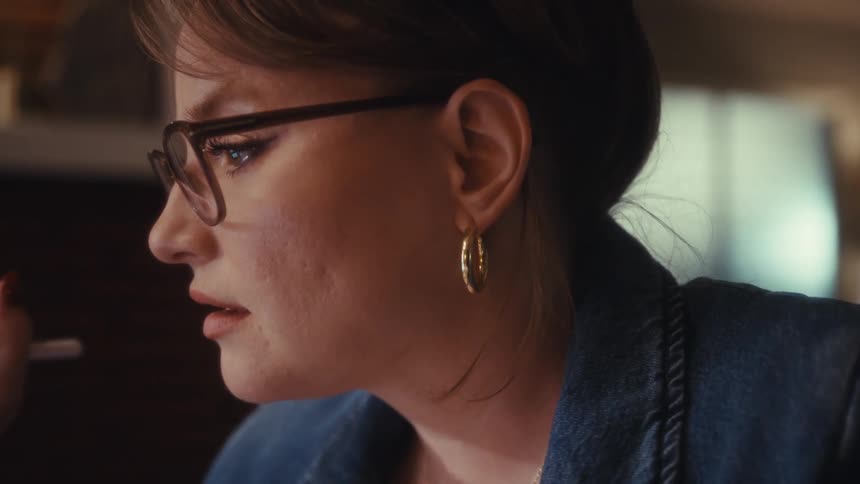 A middleaged woman sits in an office wearing glasses holding a pen while engaged in a conference call