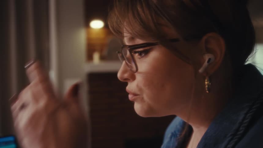 A young woman wearing glasses a wireless earbud a gold hoop earring and a denim jacket looking intently at a laptop screen while adjust her hair all within a dimly lit indoor setting