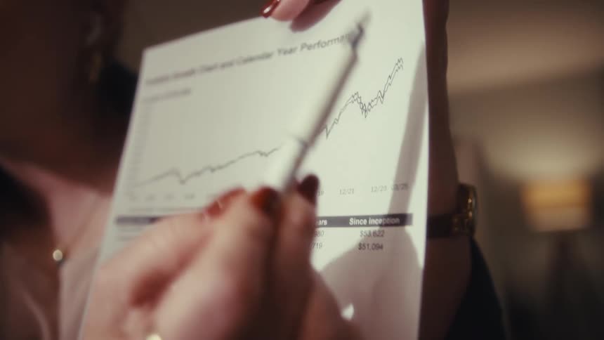 This video is a closeup of a persons hands holding a financial document titled portfolio growth chart and calendar year performance one hand with red nail polish and rings uses a pen to point to an upwardtrending line on the investment performance