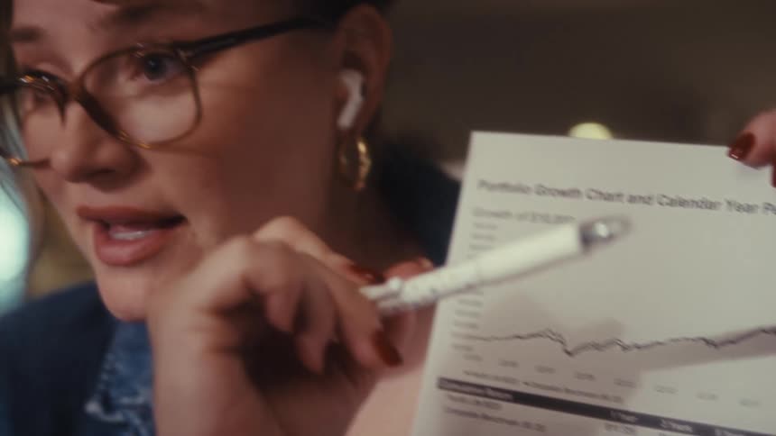 This clip features a woman with glasses holding up a financial report titled portfolio growth chart and calendar year performanceshe is smiling and nodding seemingly explaining the chart to someone offcamera possibly in a remote meeting while us