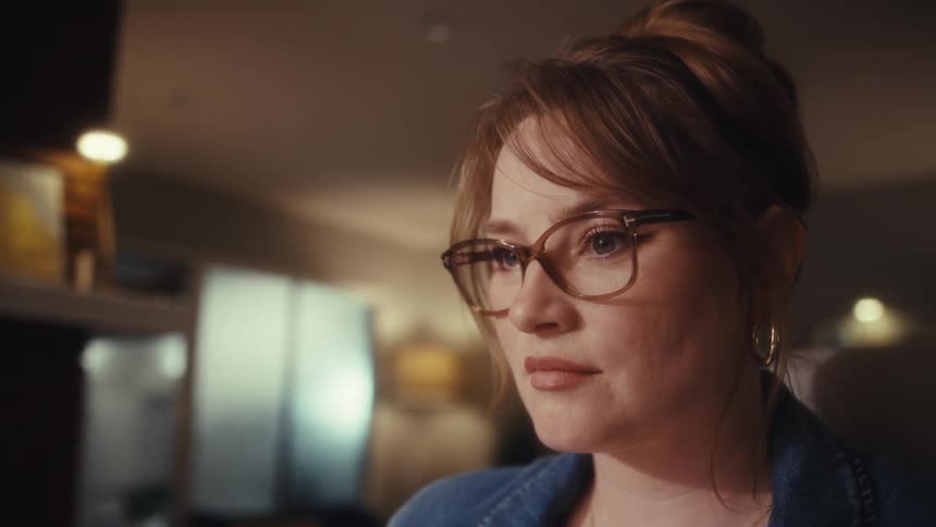 Woman with tiedup hair glasses and denim jacket focused in cozy indoor setting with soft lighting