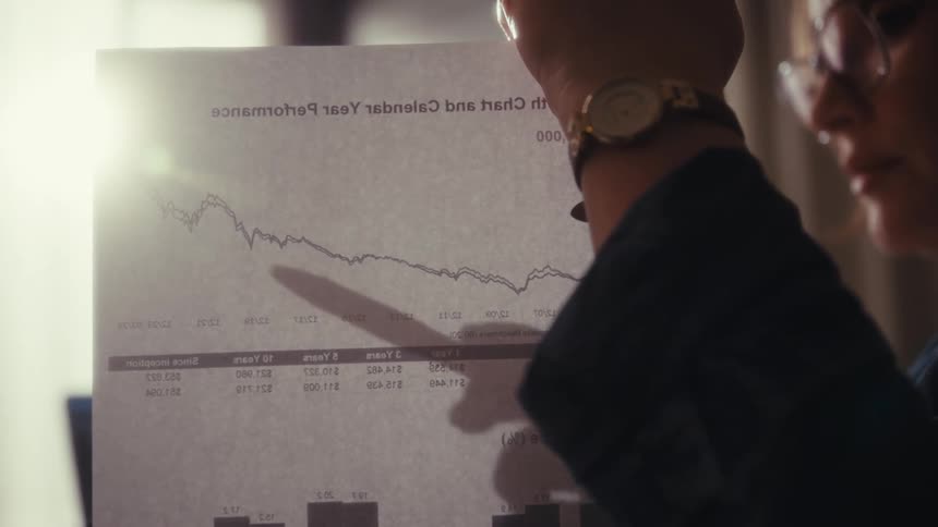 A woman is using a pen to point to a graph on a sheet of paper as she talks in an office