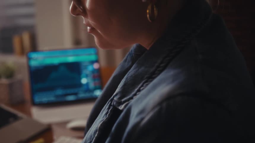 A woman with dark hair sits at a desk writing and reviewing a stock market chart on laptops displaying green and red data
