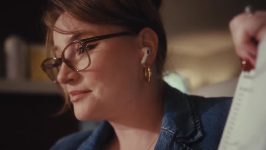 A woman in glasses and a denim jacket holds a paper with a declining chart talks into a wireless earbud and then smiles in the warm interior light