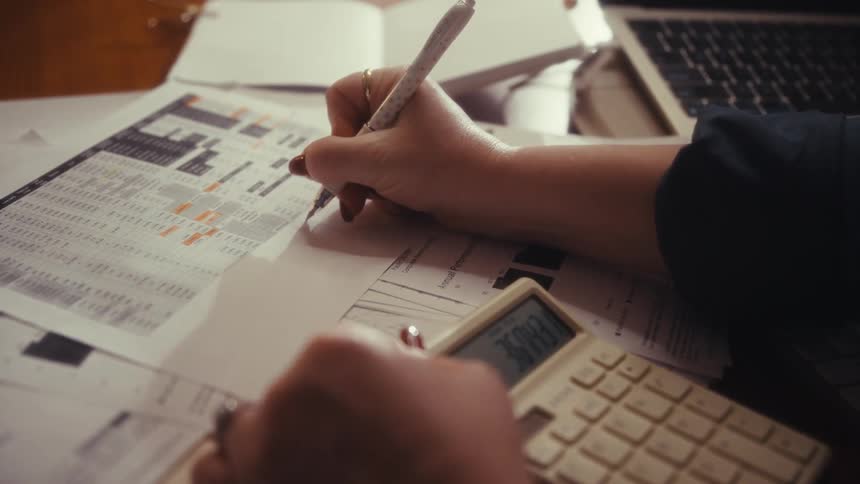 A close view of a persons hands using a calculator and writing on financial documents with a pen with a keyboard and laptop in the background