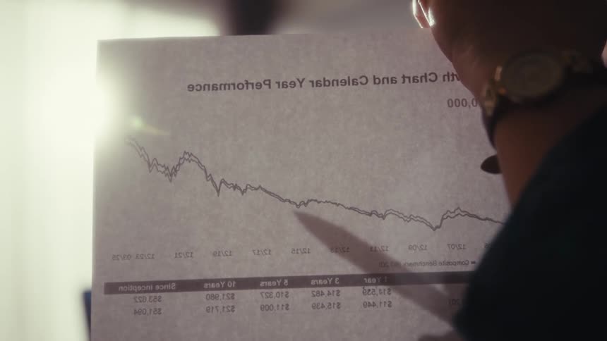 A woman is using a pen to point at a graph on a sheet of paper as she talks to someone in an office