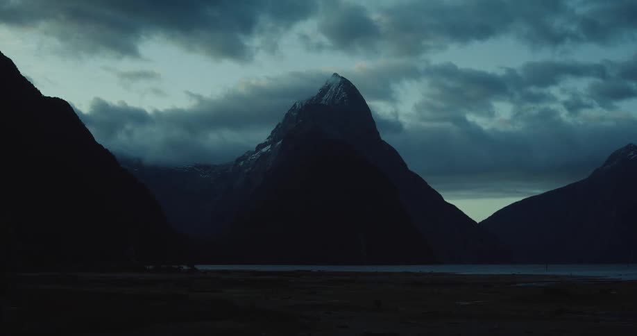 A dark mountain peak rises above a still lake shrouded in clouds under a twilight sky