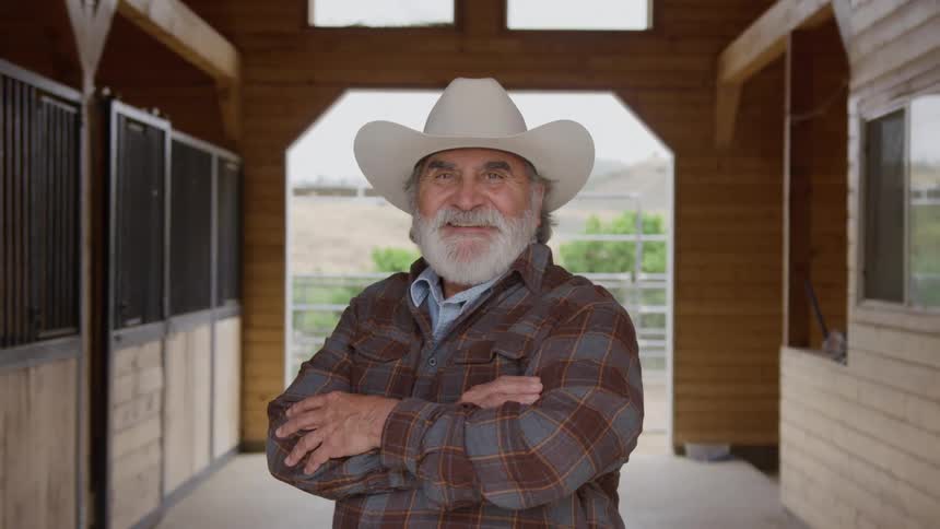 A smiling man in a cowboy hat stands with arms crossed inside a wooden barn