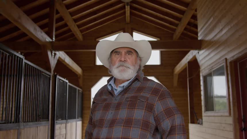 Graybearded man wears white hat and plaid shirt in wooden stable structure