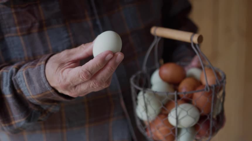 An elderly man is holding an egg in one hand and a basket of eggs in the other