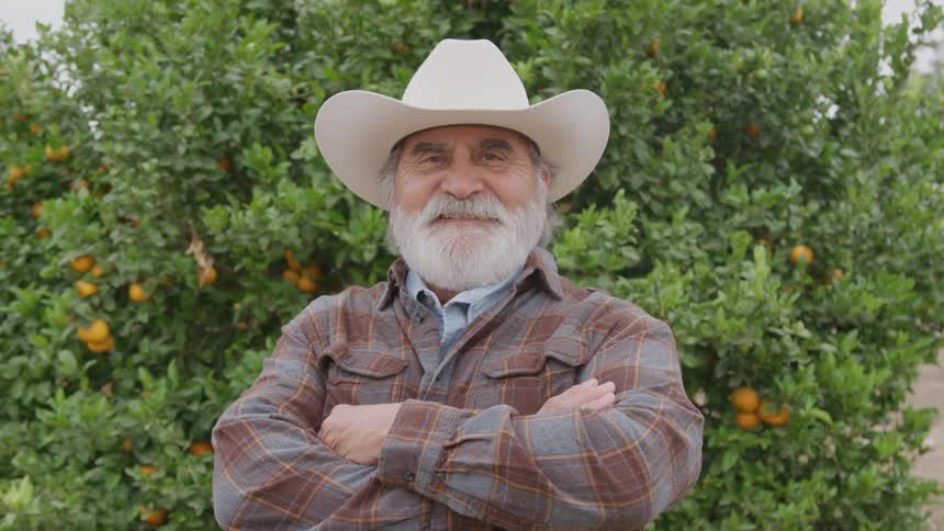 An older man with a white beard and a cowboy hat wearing a plaid shirt and standing with his arms crossed as he smiles directly at the camera in front of a heavily laden orange tree