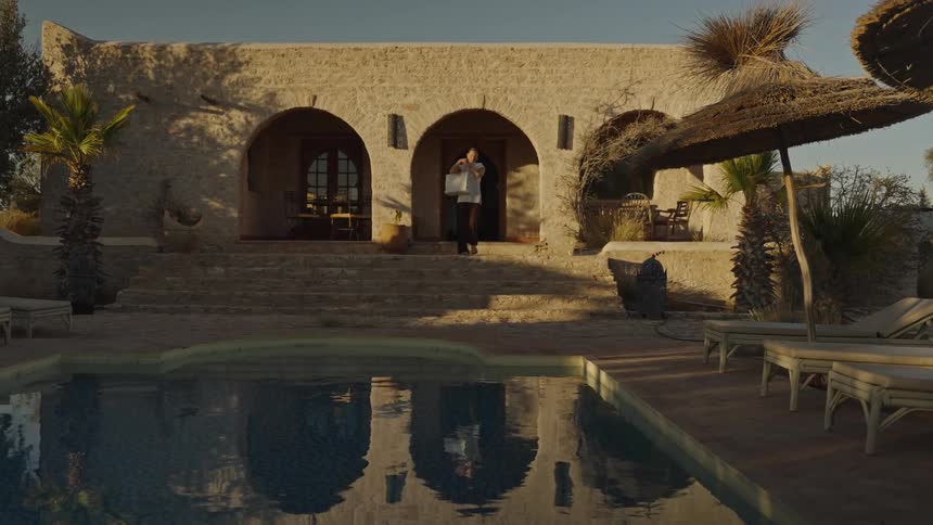 This video is a wide shot of a mediterranean or desertstyle villa with a swimming pool in the foregrounda person wearing a white dress and carrying a large bag walks out from one of the arched doorways of the stone building towards the pool area whi